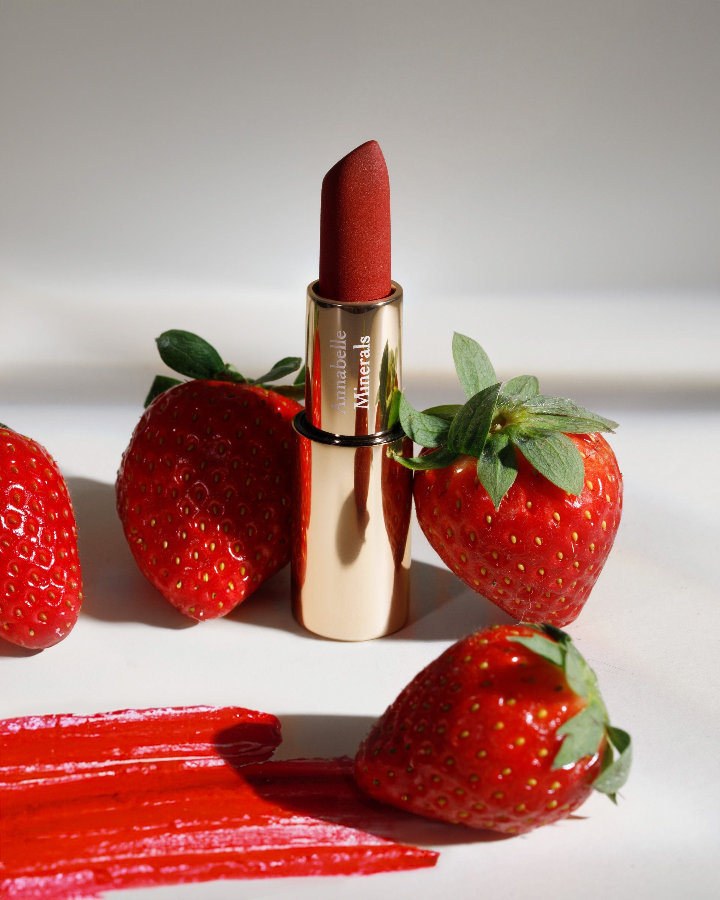 Pomadka do ust w soczyście czerwonym odcieniu Strawberry Annabelle Minerals w otoczeniu truskawek ze swatchem koloru