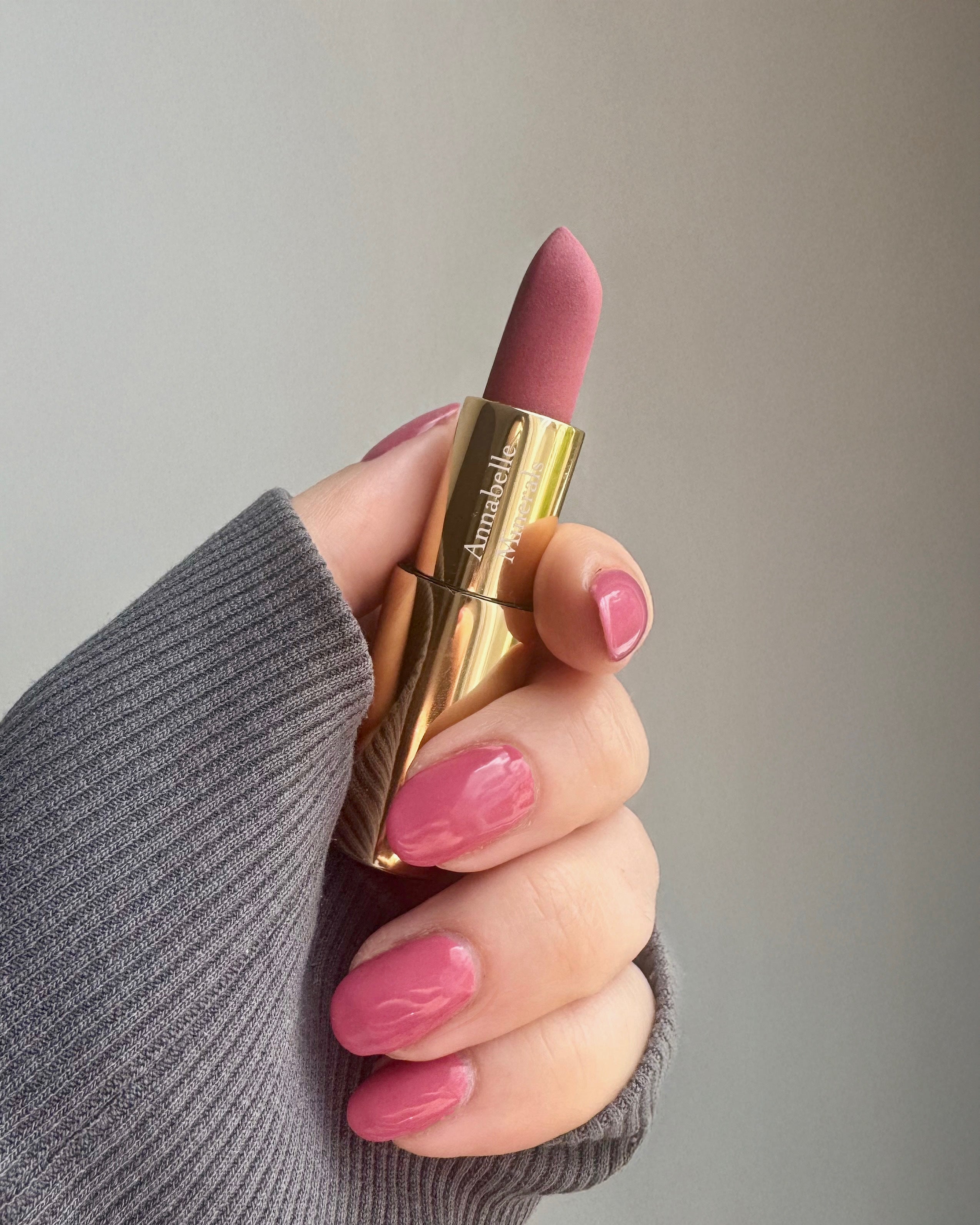 Różowa szminka Pink Apple Annabelle Minerals w złotym opakowaniu trzymana w dłoni z różowym manicure, w tle szary sweter