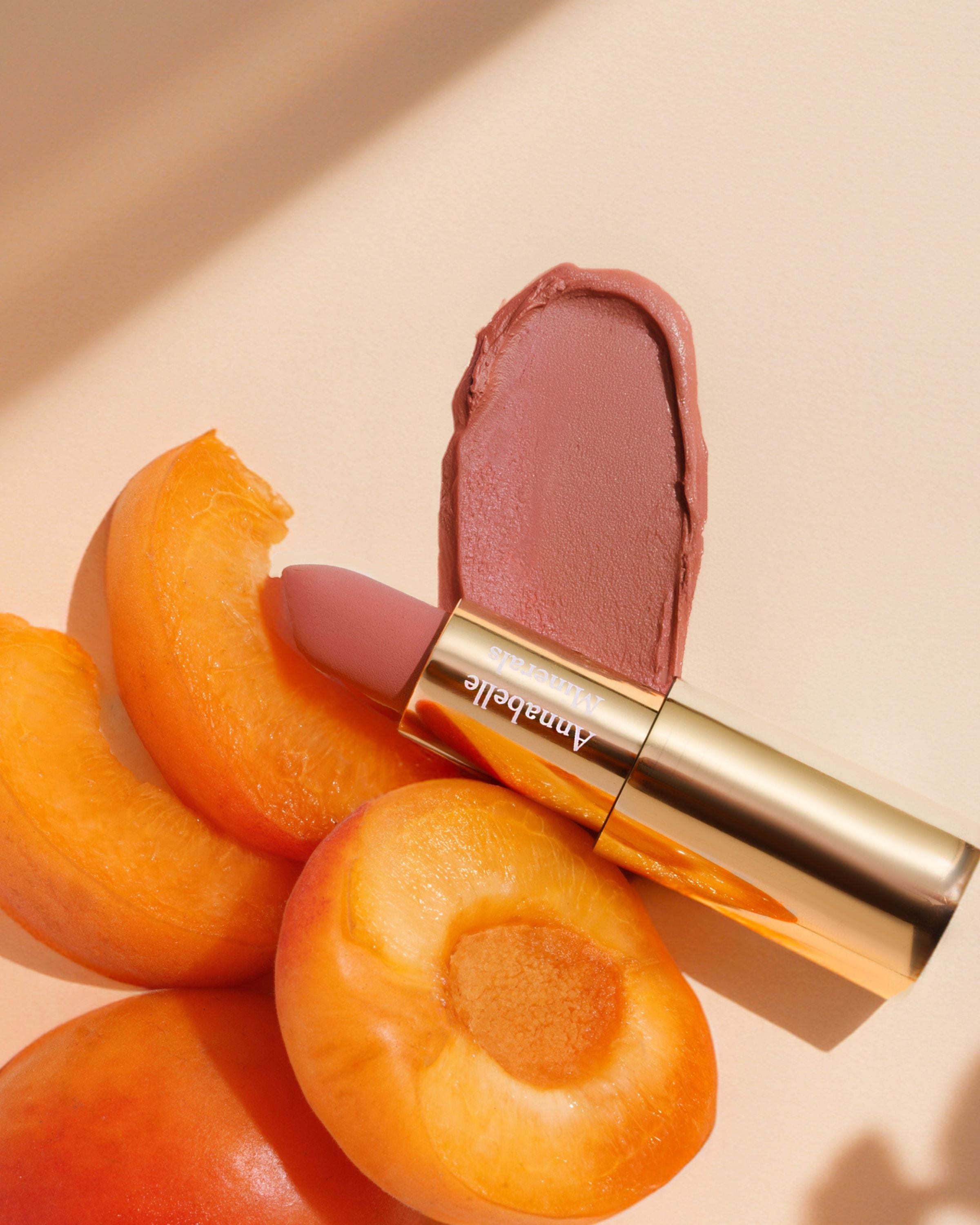 Pomadka do ust w odcieniu Apricot Annabelle Minerals w otoczeniu owoców moreli i swatcha koloru